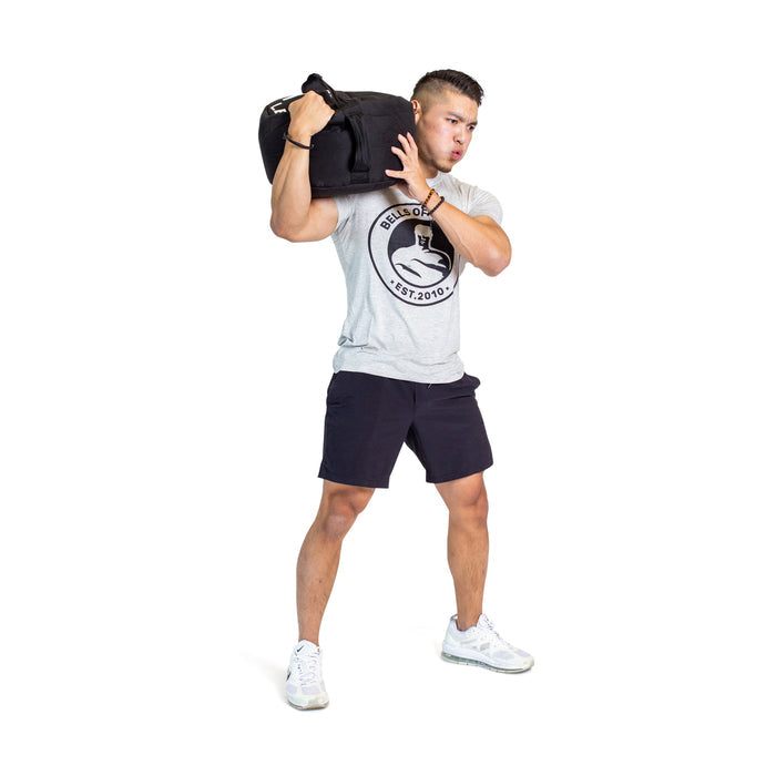 A man in athletic wear poses mid-exercise with a Bells of Steel Fitness Sandbag on his right shoulder, emphasizing the challenging nature of sandbag workouts against a plain white background.