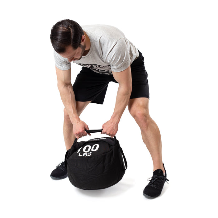 A man in a grey t-shirt and black shorts lifts a Bells of Steel Fitness Sandbag labeled "100 LBS" with both hands against a white background, highlighting the challenge and intensity of sandbag training.