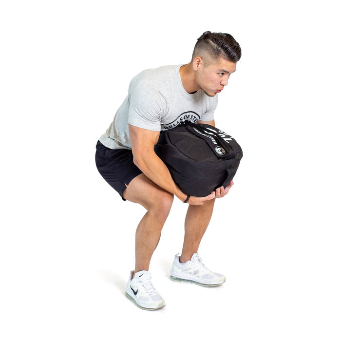 A man in a grey t-shirt, black shorts, and white sneakers demonstrates a Bells of Steel Fitness Sandbags workout by squatting and lifting the sandbag with both hands against a white background.