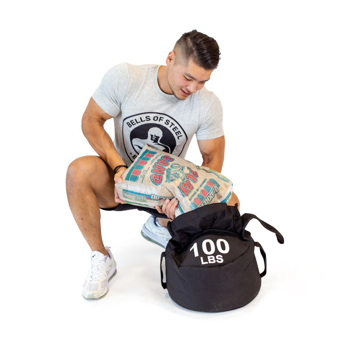 A man in athletic wear pours gravel into a Bells of Steel Fitness Sandbag labeled "100 LBS" against a white background, perfect for sandbag workouts.