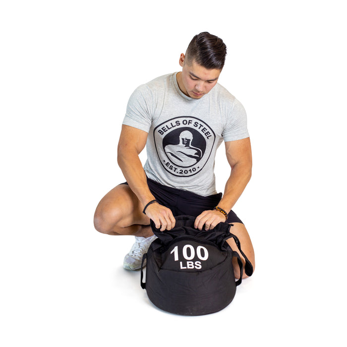 A person in a gray Bells of Steel t-shirt kneels on one knee, adjusting a black Bells of Steel Fitness Sandbag labeled "100 LBS" against a white background—ideal for workouts and made with durable Condura construction.