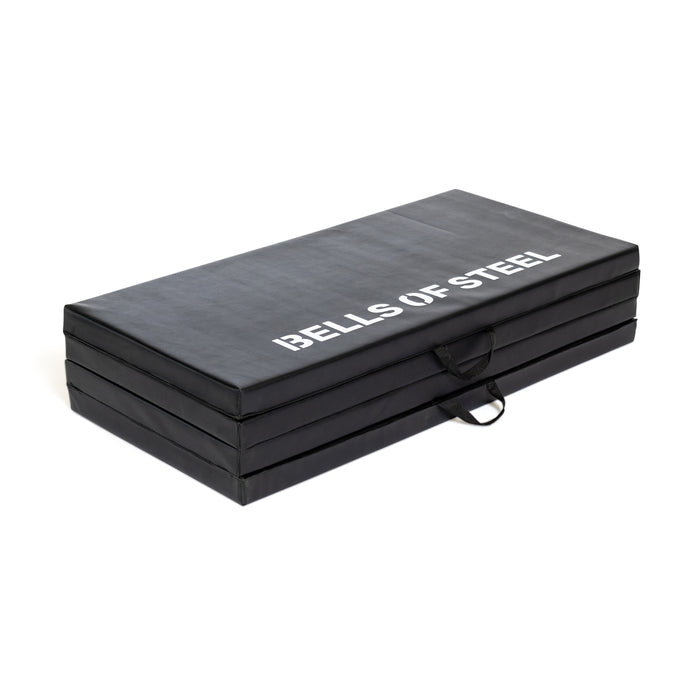The Bells of Steel Four Fold Mat is a black, foldable plush foam exercise mat with white branding on top. It features four stackable sections and carrying handles for portability.