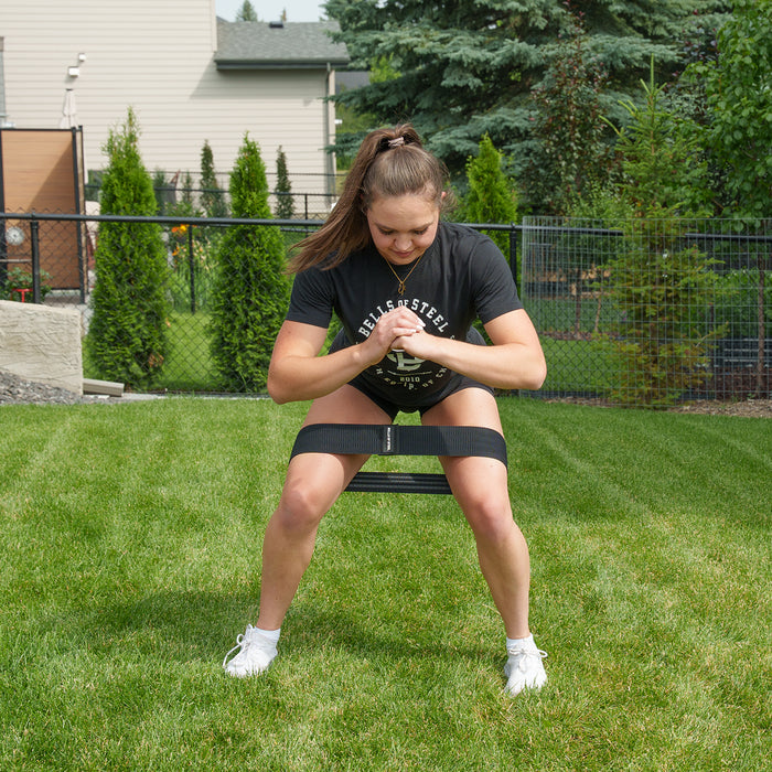 A person wearing a black shirt and shorts exercises outdoors with the Bells of Steel Glute Loop resistance band positioned above the knees, squatting on grass with hands clasped in front, in a fenced backyard for an effective booty workout.