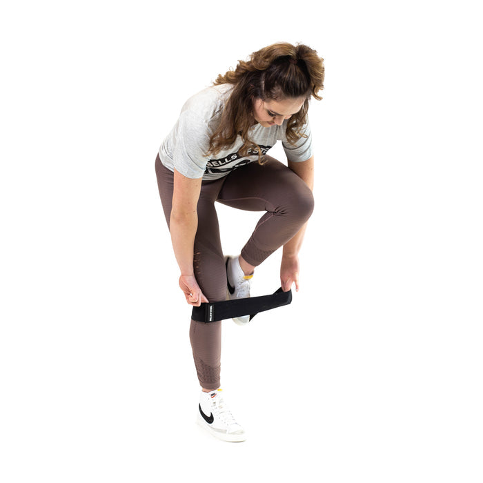 A woman in athletic wear balances on one leg, using the Bells of Steel Glute Loop around her foot for a booty workout against a white background.