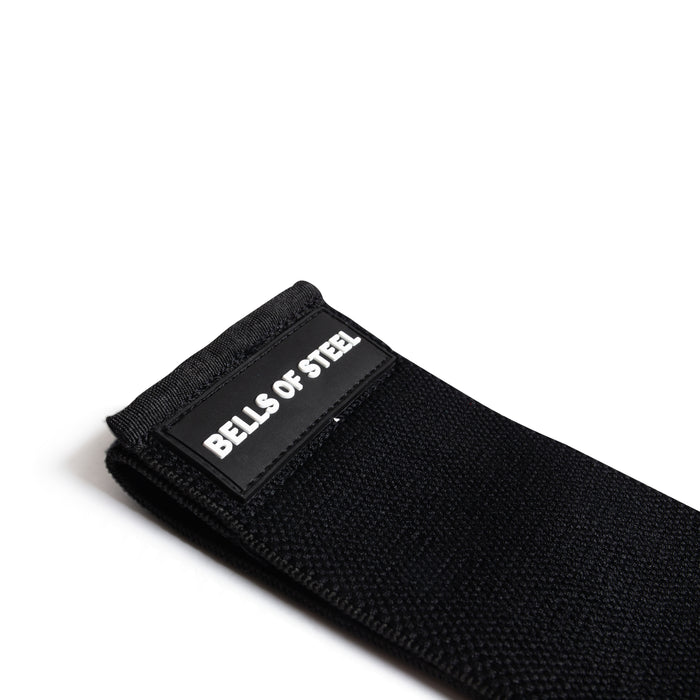 A close-up of the Bells of Steel Glute Loop—black fabric strap with a white "BELLS OF STEEL" label—set against a plain white background, ideal for booty workouts.