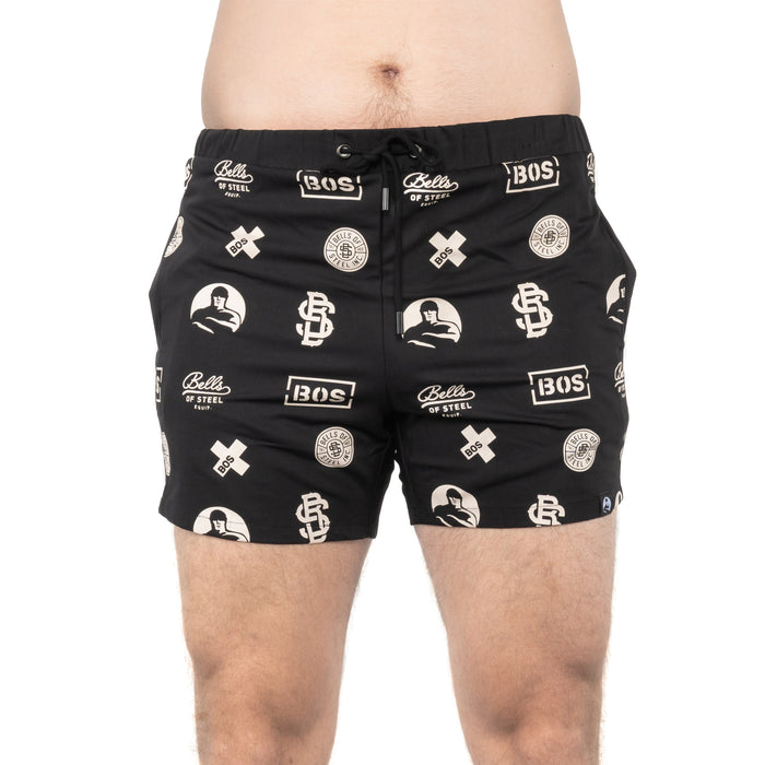 A person wears Bells of Steel Gym Shorts featuring white logos, text, and graphic designs while standing against a white background.
