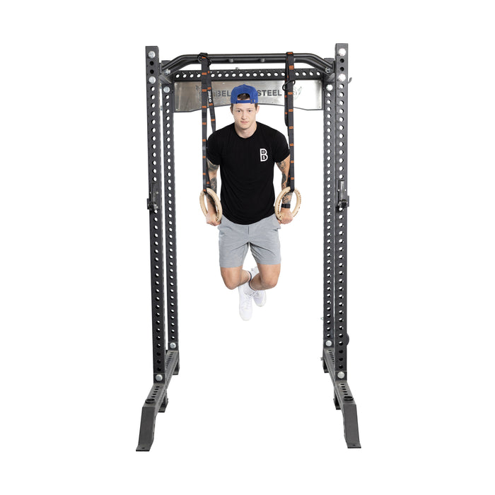 Wearing a blue cap, black T-shirt, and gray shorts, a person performs a dip using Bells of Steel Gymnastics Rings attached to a metal rack against a plain white background.