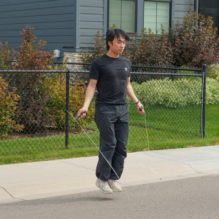 Wearing black athletic clothing, a person is skipping rope on a sidewalk in front of a house with a fenced yard and green bushes, engaging in cardio training with the Bells of Steel Jump Rope, which features ergonomic handles and an adjustable design.