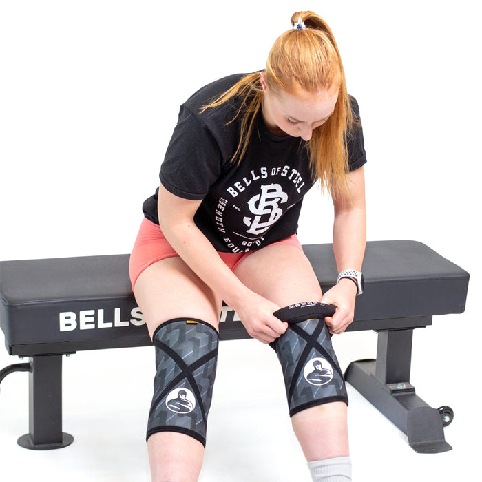 A woman with red hair tied back sits on a workout bench in pink shorts and a Bells of Steel shirt, adjusting Bells of Steel Knee Sleeves on both legs. The background is plain white.