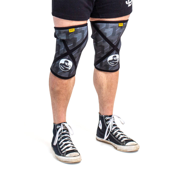 A person wears Bells of Steel Knee Sleeves featuring a white graphic, paired with black-and-white high-top sneakers. Only their legs, knees, and feet are shown against a white background.