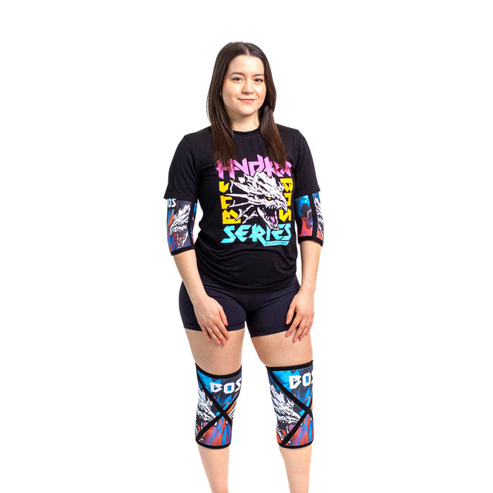 A woman faces forward, smiling against a white background, wearing Bells of Steel Knee Sleeves with bold patterns and "BOSS" on the knees for squat support, along with a black graphic T-shirt and black shorts.