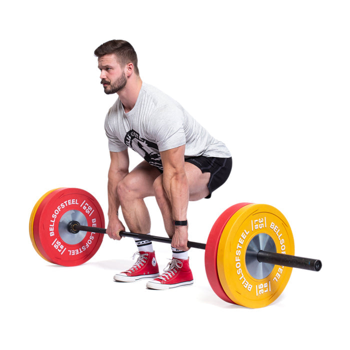 A man in a gray t-shirt, black shorts, and red shoes prepares to lift a barbell loaded with Bells of Steel Boltless LB Competition Bumper Plates that meet IWF specs, against a white background, capturing Olympic lifting spirit.