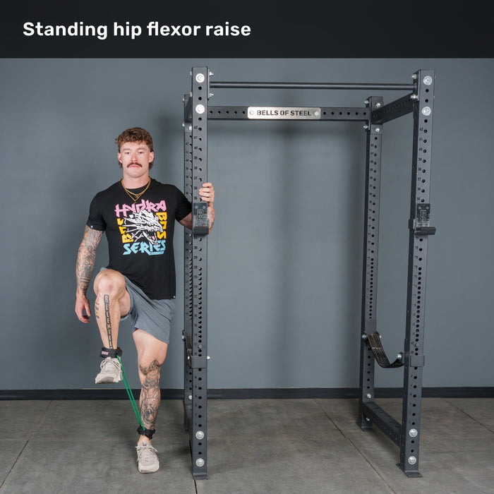 A man performs a standing hip flexor raise with Bells of Steel USA Leg Cords, holding onto a metal rack for balance. He lifts one knee toward his chest while keeping the other leg straight.