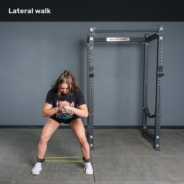 A woman in athletic wear performs a lateral walk using Bells of Steel USA Leg Cords around her legs in a gym with a squat rack behind her.