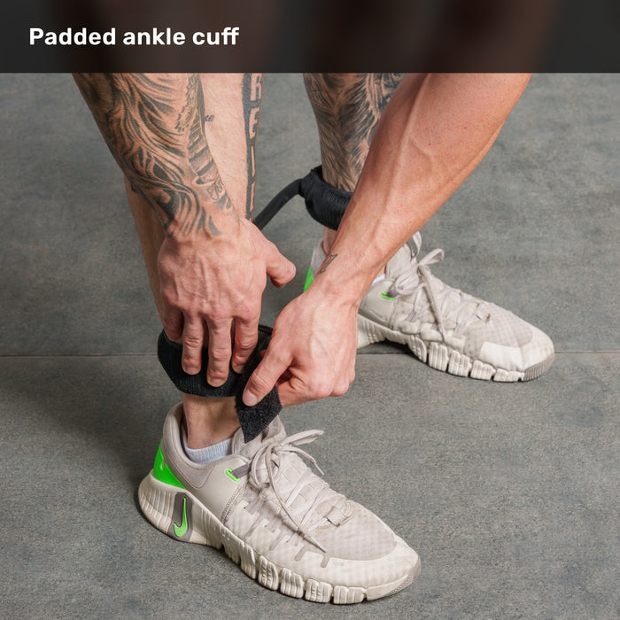 A person with tattoos fastens a padded ankle cuff for the Bells of Steel USA Leg Cords resistance bands over their left ankle while wearing light gray sneakers with green accents on a gray floor.