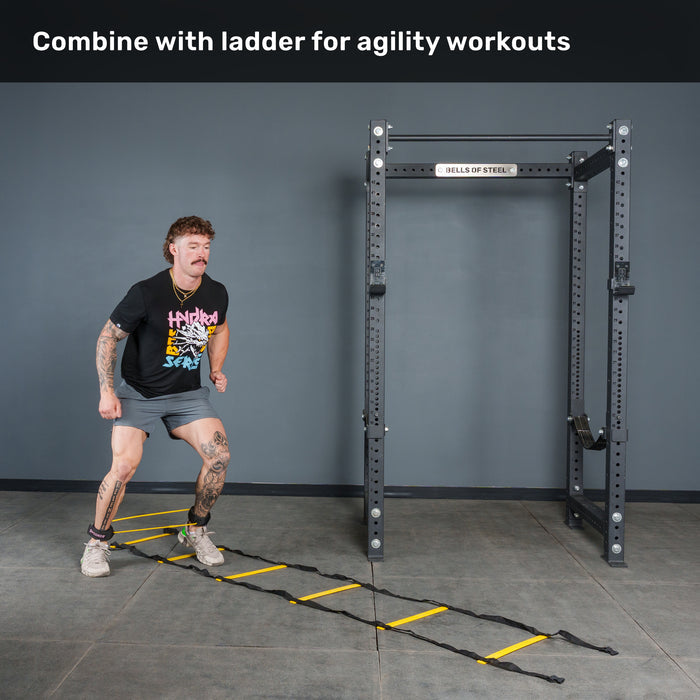 A man in athletic wear exercises on an agility ladder in a gym next to a black squat rack. Text above reads, “Combine with ladder for agility workouts using Bells of Steel USA Leg Cords.”.
