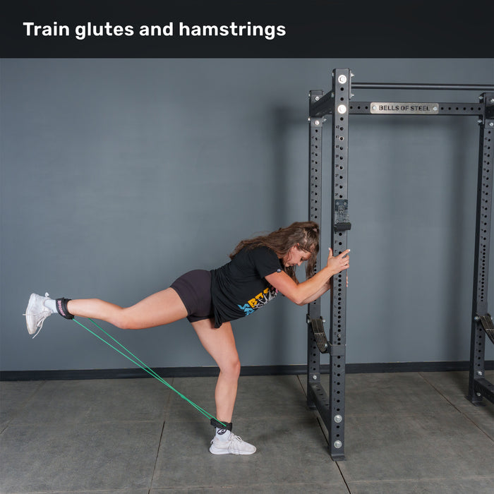 Using the Bells of Steel USA Leg Cords, a person exercises their glutes and hamstrings by extending one leg back with the resistance band attached to their ankle and a power rack, while holding onto the rack for support.