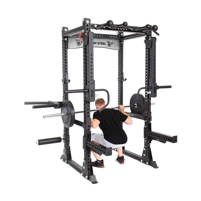 Wearing a black T-shirt and gray shorts, a person squats with a barbell in a Bells of Steel Manticore Flat Foot Components power rack, equipped with safety bars and various attachments. The sturdy black rack features the brand's logo prominently at the top.