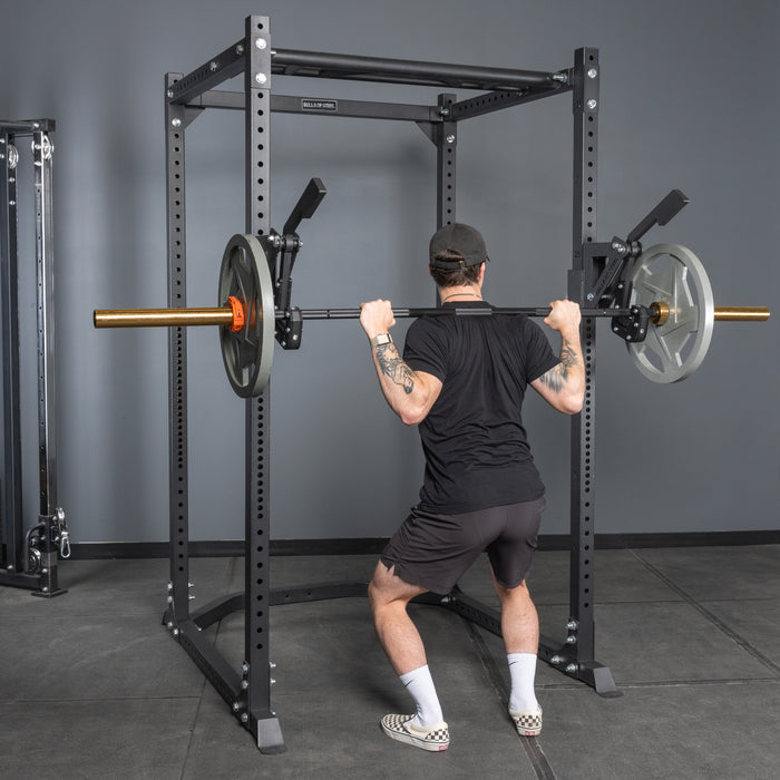 A person in a black outfit is performing a barbell squat inside a gym power rack equipped with the Bells of Steel Monolift Rack Attachment, using weighted plates and facing away from the camera.