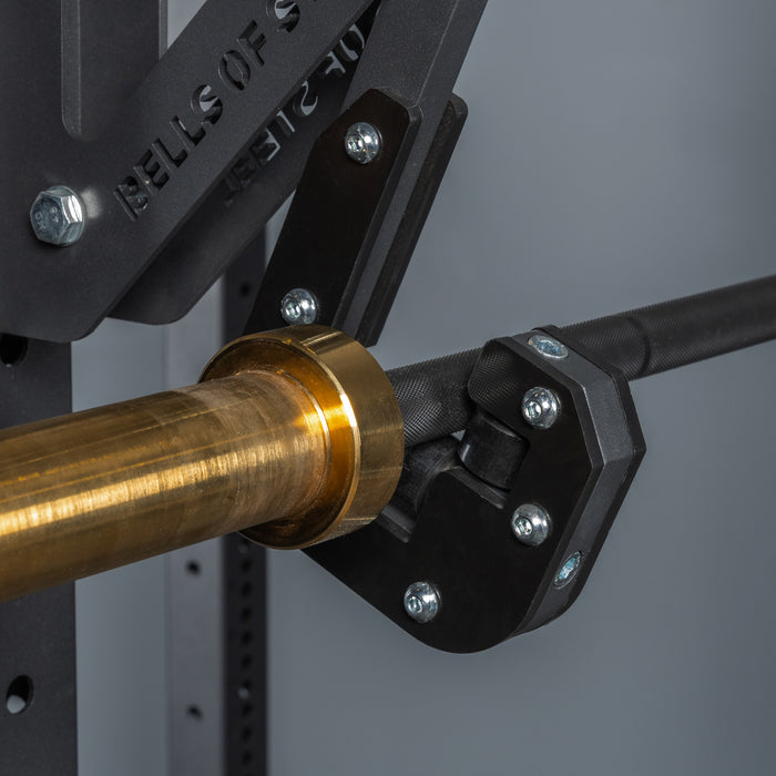 Close-up of a barbell with a gold sleeve secured by the Bells of Steel Monolift Rack Attachment, a sturdy, adjustable, black metal holder bolted to the squat rack for efficient, space-saving support.