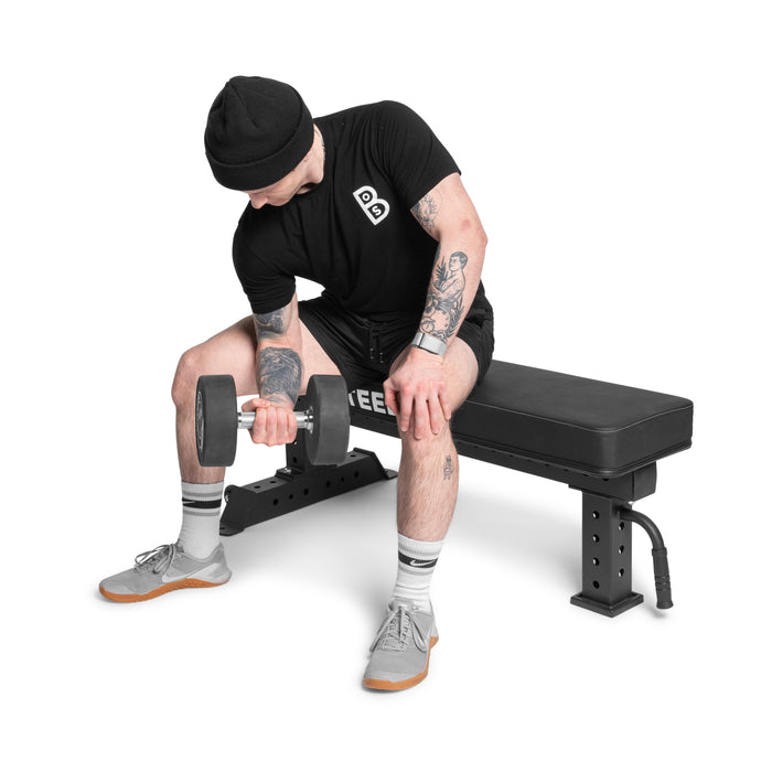 A person wearing a black t-shirt, shorts, beanie, and sneakers sits on the Bells of Steel Powerlifting Flat Bench, performing a seated bicep curl with a dumbbell.