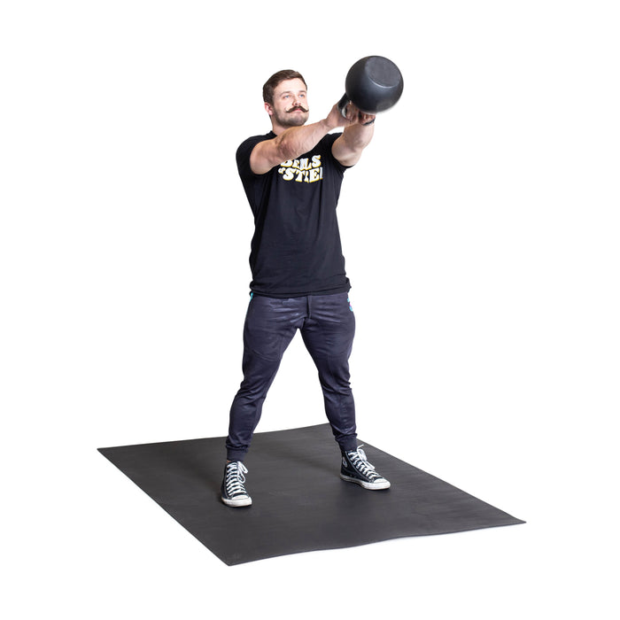 A man stands on the Bells of Steel Premium Rubber Flooring 4' x 6', lifting a kettlebell at chest height. He wears a black T-shirt, dark pants, and sneakers against a plain white background.
