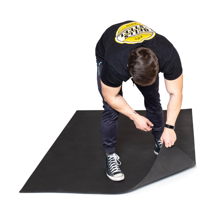 A person in black attire lifts the corner of a Bells of Steel Premium Rubber Flooring 4' x 6' mat, highlighting the gym’s durable recycled rubber surface.