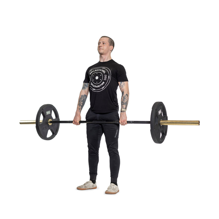A person stands holding a barbell with ergonomic handles and Bells of Steel Rubber Coated Iron Weight Plates, dressed in a black t-shirt and pants against a plain white background.