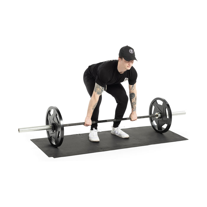 Wearing black athletic wear and white sneakers, a person deadlifts with both hands on a barbell atop Bells of Steel Rubber Flooring Puzzle Gym Mat 24" x 24" (Set of 8), ensuring optimal floor protection.