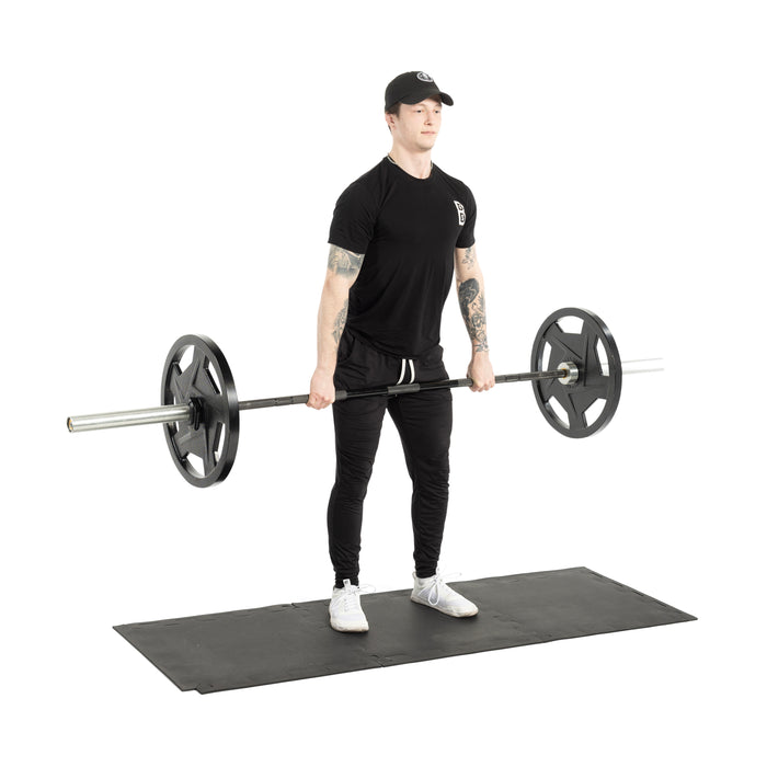 A person in athletic gear stands on the Bells of Steel Rubber Flooring Puzzle Gym Mat 24" x 24" (Set of 8), holding a barbell at thigh level. The clean white background highlights the gym floor protection these mats provide.