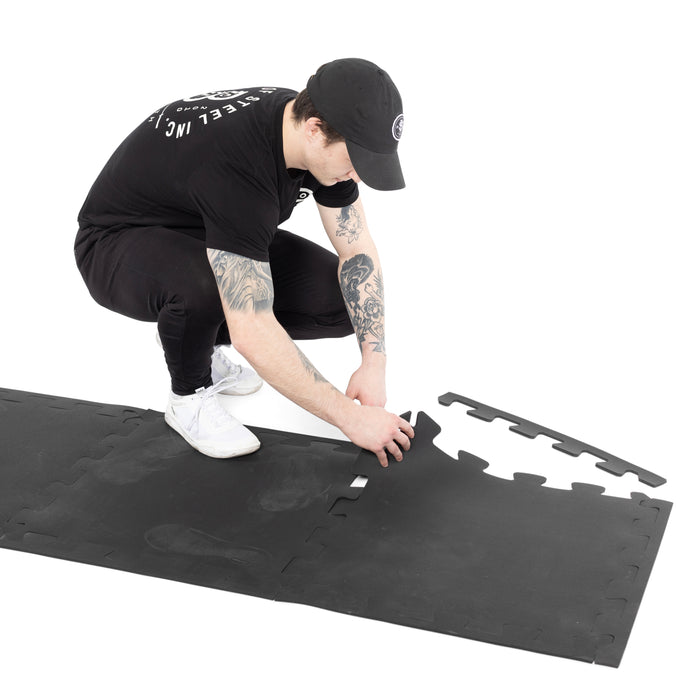 A person in black clothing and a cap kneels on a white background, connecting two Bells of Steel Rubber Flooring Puzzle Gym Mat 24" x 24" (Set of 8) pieces—perfect for gym floor protection.