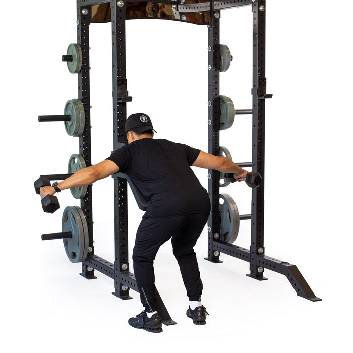 Wearing black workout clothes, a person does a bent-over dumbbell raise in front of a squat rack with the Bells of Steel Seal Row Pad Rack Attachment and weight plates.