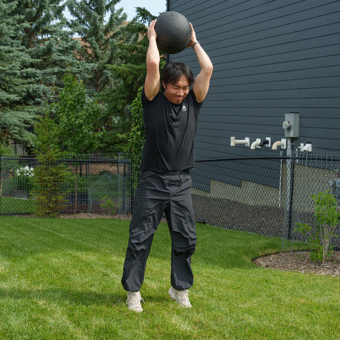 A person dressed in black athletic wear stands on grass, holding a Bells of Steel Slam Ball overhead and preparing to slam it down, demonstrating explosive power. In the background are trees, a fenced yard, and a dark building.