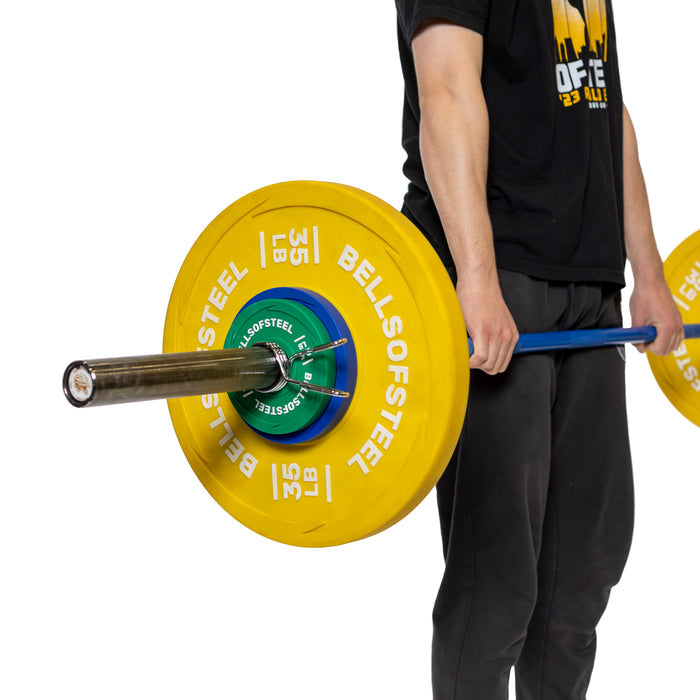 A person in black clothing lifts a barbell with polished chrome Olympic sleeves, using Bells of Steel yellow weight plates marked "35 LB" and secured by Spring Collars. The weights are blue and green in the center.