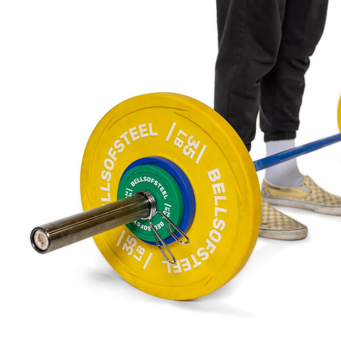 A close-up of a barbell with vibrant yellow and green weight plates, marked "Bells of Steel," secured by Spring Collars. The polished chrome gleams under the light, while checkered slip-on shoes await in the background.