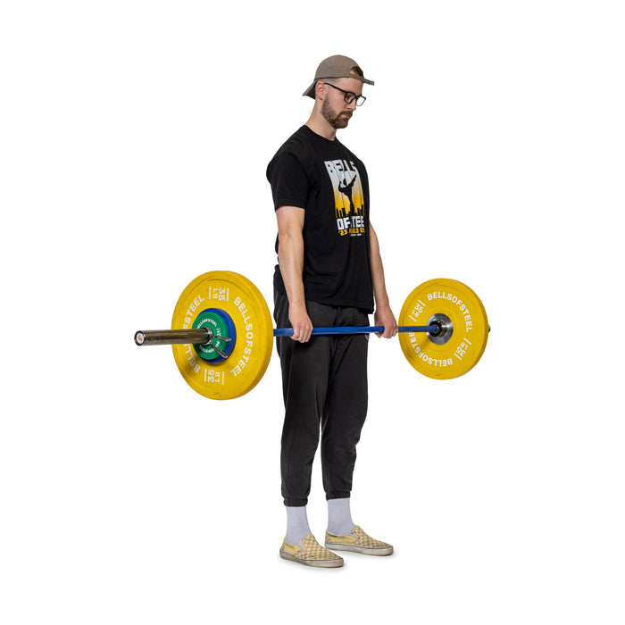 A man confidently holds a barbell with Bells of Steel Spring Collars securing yellow plates on the Olympic barbell sleeves. He stands against a white background in casual clothing, including a cap, glasses, black t-shirt, and checkered shoes.