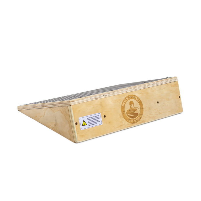 The Bells of Steel Squat Wedge Board features a wood build, metal grip surface, safety warning label, and a circular logo with figure and text. It's designed to boost ankle mobility and squat depth during stretching or exercise.