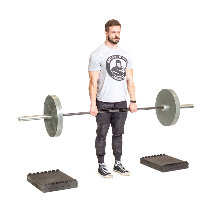 A man in a gray t-shirt and black pants is performing deficit deadlifts with a barbell on Bells of Steel Stackable Pull Blocks, positioned between two black rubber mats against a white background.