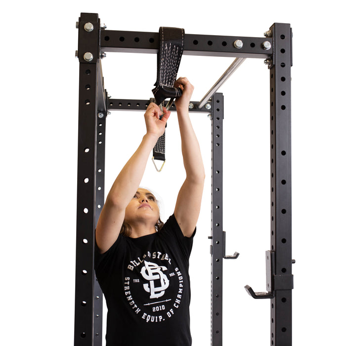 In a sleek home gym, a person in a black T-shirt expertly attaches Bells of Steel's Suspension Spotter Straps And Bodyweight System to the pull-up bar on a sturdy black metal power rack for invigorating suspension training.