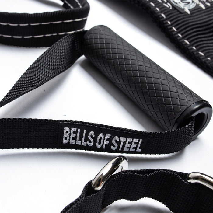 Close-up of a textured black exercise handle from Bells of Steel's Suspension Spotter Straps and Bodyweight System, perfect for home gym setups. The handle is attached to a strap with metal rings and stitching details, labeled "Bells of Steel," resting on a white surface.