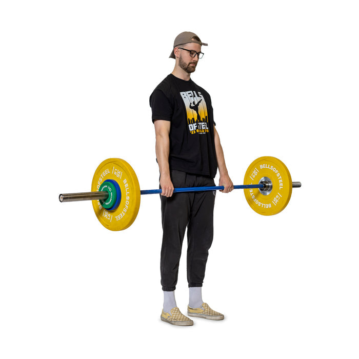 A man in glasses, a cap, T-shirt, pants, and slip-on shoes holds a barbell loaded with Bells of Steel Urethane-Coated Change Plates, preparing to lift against a plain white background.