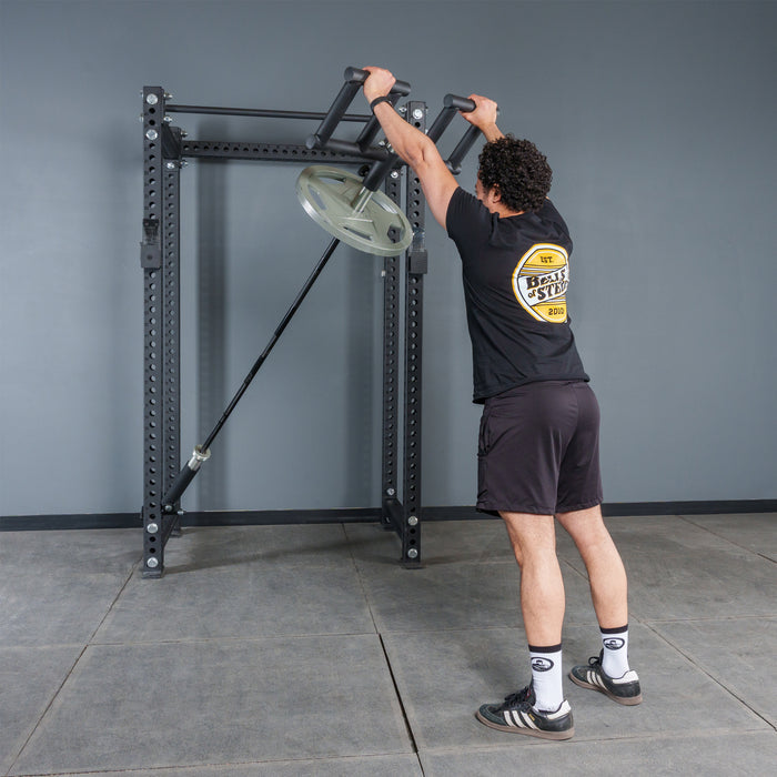 A person is using the Bells of Steel Viking Press Attachment with an Olympic bar and weight plate in a gym with gray walls and flooring, wearing a black T-shirt, shorts, and sneakers.