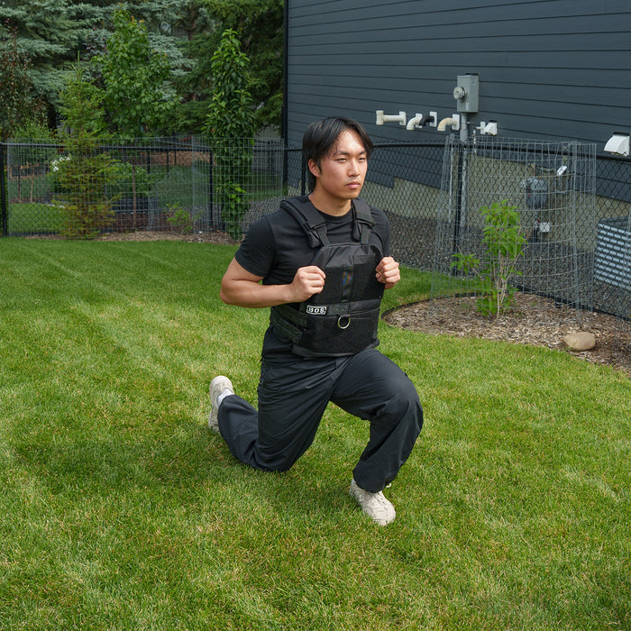 A person wearing the Bells of Steel USA Weighted Vest and Plates over black workout clothes performs a lunge on green grass in a fenced backyard beside a modern dark house.