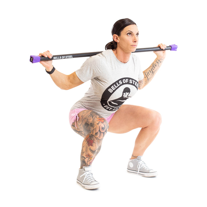 A person strength trains, squatting with Weighted Workout Body Bars by Bells of Steel. They sport a Bells of Steel t-shirt, pink shorts, and gray sneakers. Tattoos embellish one leg as they pose against the stark white background, highlighting their fitness style.