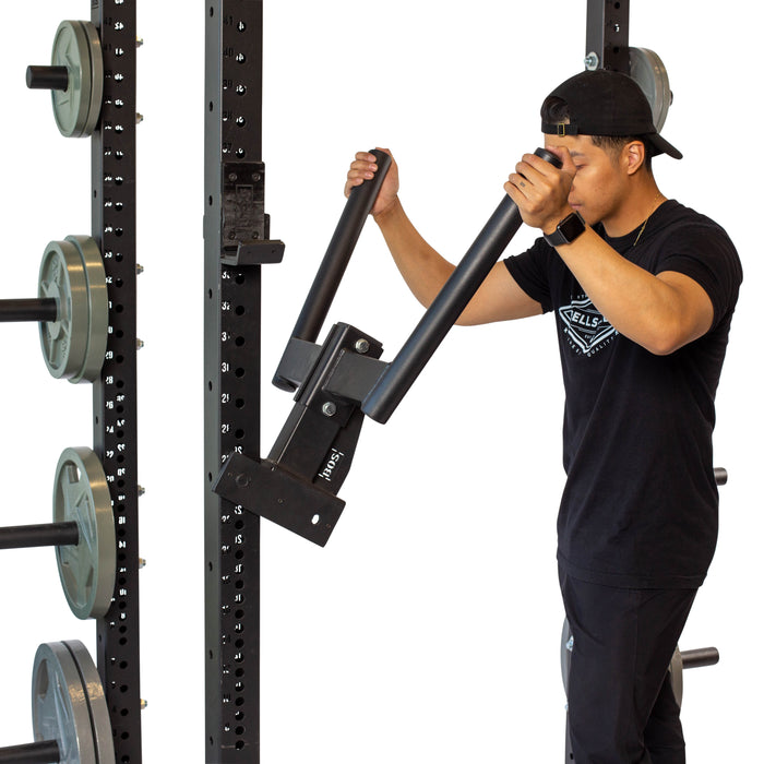Wearing a black shirt and cap, someone confidently uses the Bells of Steel Y Dip Bar Rack Attachment on a squat rack with a 500lb capacity. Surrounded by weight plates, they grip the handles while standing in profile.